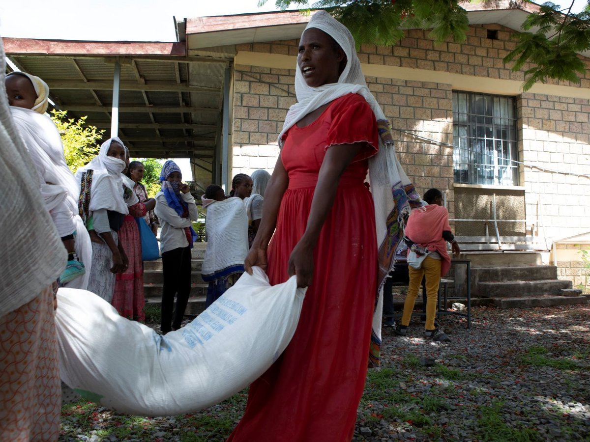 Eine Frau in rotem Kleid und weißem Kopftuch trägt einen großen Hilfssack, mit anderen Frauen und Kindern im Hintergrund.