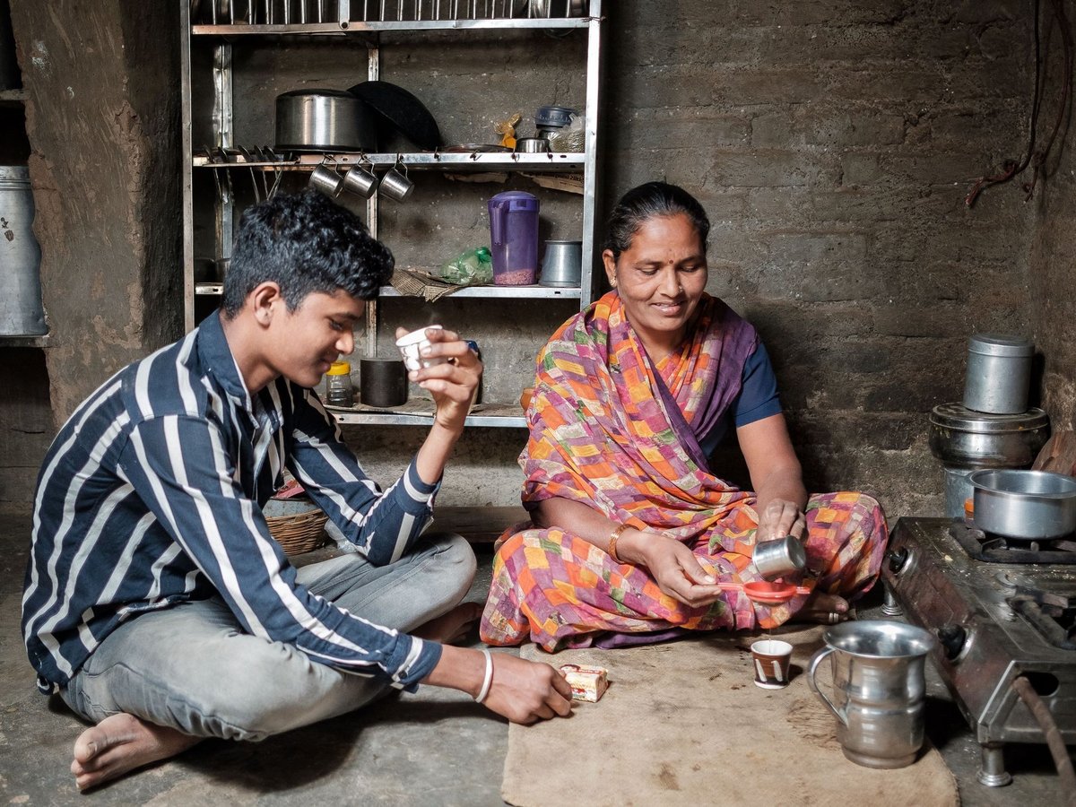 Eine indische Frau gießt Tee für einen lächelnden jungen Mann in einer einfachen Küche mit Metallregalen und Gasherd.