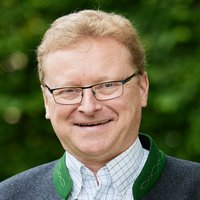 Lächelnder Mann mit Brille in einer traditionellen Trachtenjacke vor grünem Hintergrund.