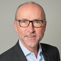 Porträt eines lächelnden Mannes mittleren Alters mit Brille, grauem Haar und Bart, im dunklen Anzug und hellblauen Hemd.