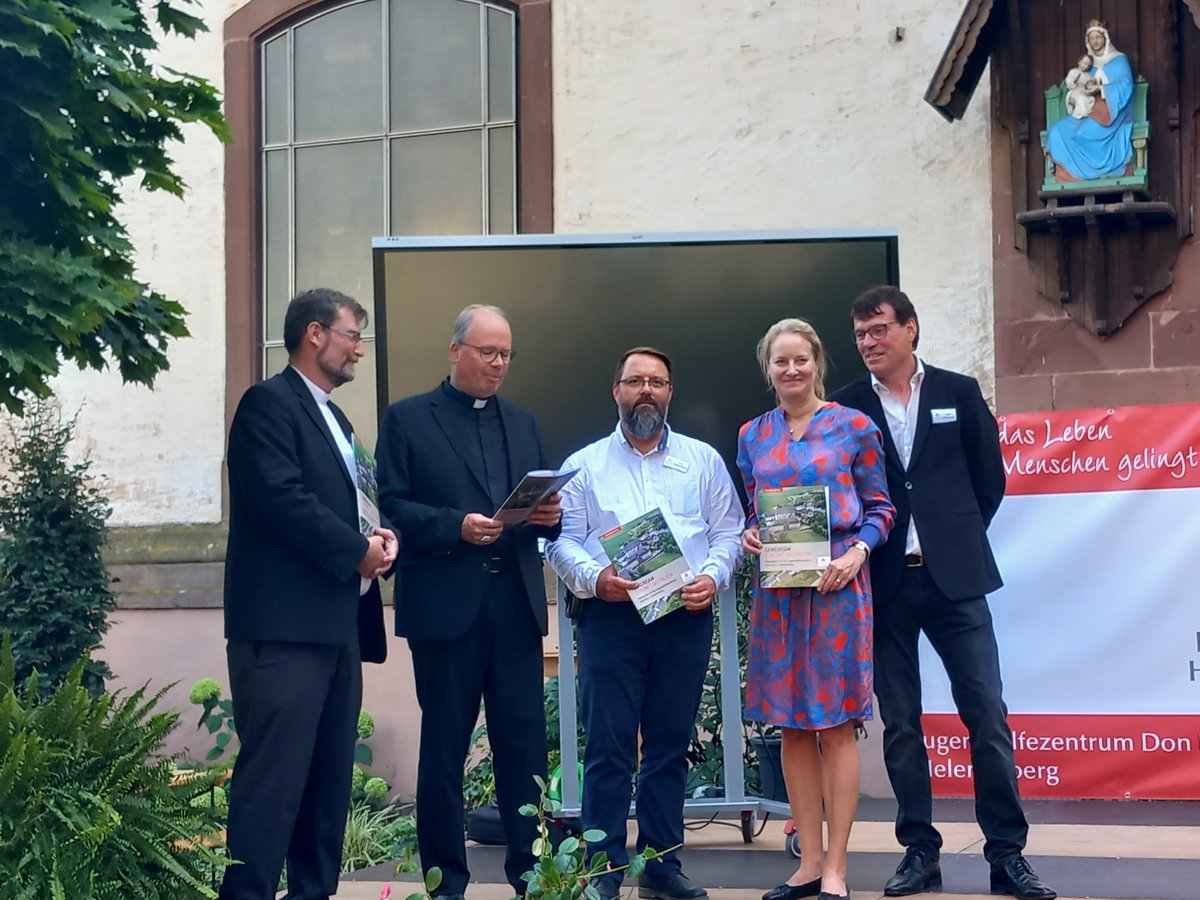 Fünf Personen, darunter Geistliche, präsentieren Broschüren vor einer Kirche mit Marienstatue und rotem Banner.