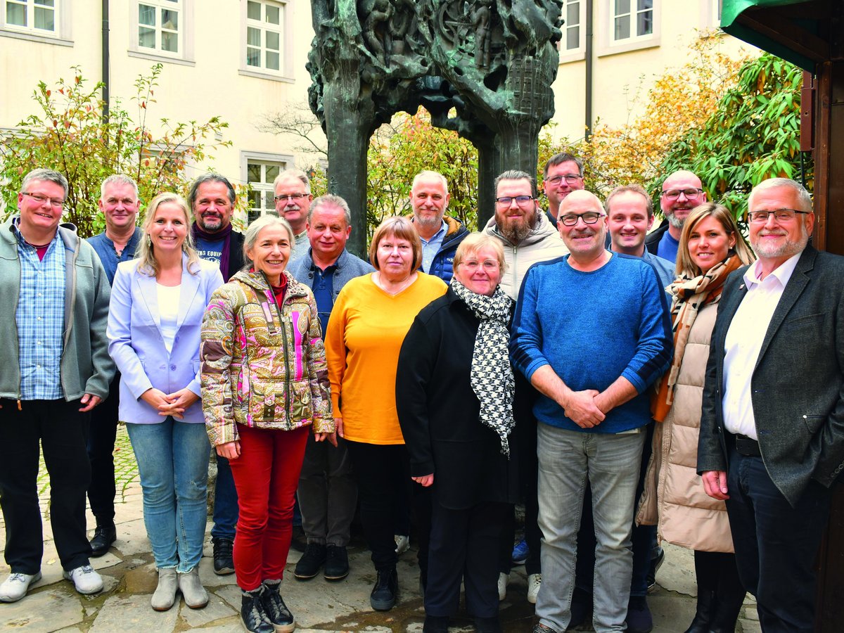Eine lächelnde Gruppe von etwa zwanzig Männern und Frauen steht im Freien vor einem Gebäude und einer großen Statue.