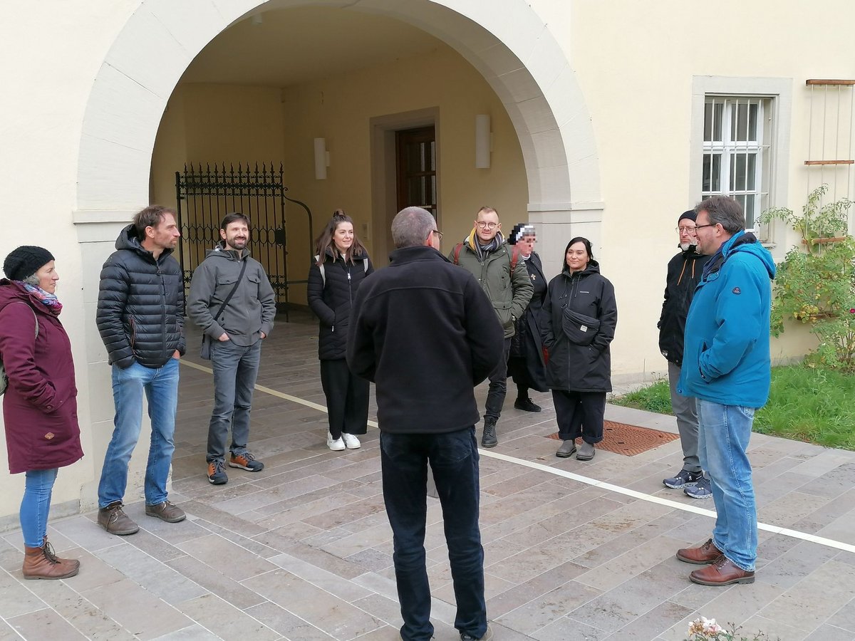 Eine Gruppe von Erwachsenen hört einem Sprecher in einem gepflasterten Außenhof vor einem Steinbau mit Torbogen zu.