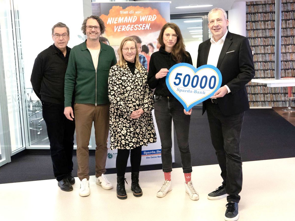 Fünf Personen, zwei Frauen und drei Männer, posieren mit einem herzförmigen Schild „50000 Sparda-Bank“ vor einem Banner „NIEMAND WIRD VERGESSEN“.
