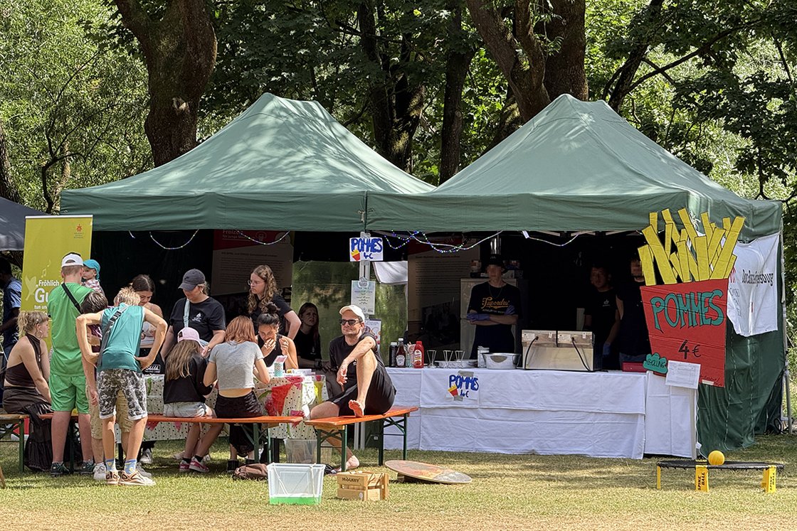 Eine Gruppe junger Leute an einem Imbissstand unter grünen Zelten im Freien, mit einem großen Pommes-Schild.