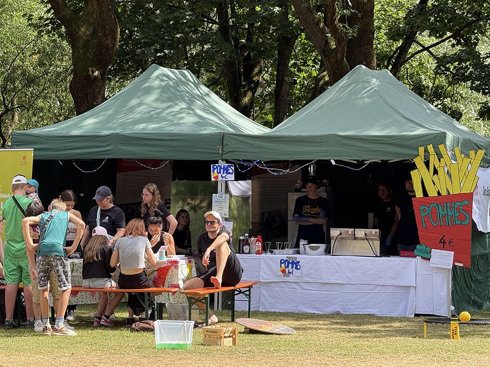 Eine Gruppe junger Leute an einem Imbissstand unter grünen Zelten im Freien, mit einem großen Pommes-Schild.