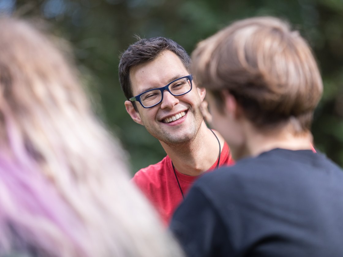 Ein lächelnder Mann mit dunklem Haar und blauer Brille in einem roten Hemd unterhält sich fröhlich mit verschwommenen Freunden im Freien.