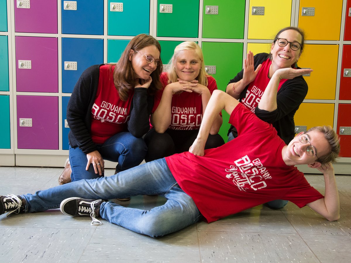 Vier lächelnde Personen, drei Frauen und ein Mann, in roten Hemden posieren spielerisch vor bunten Spinden.