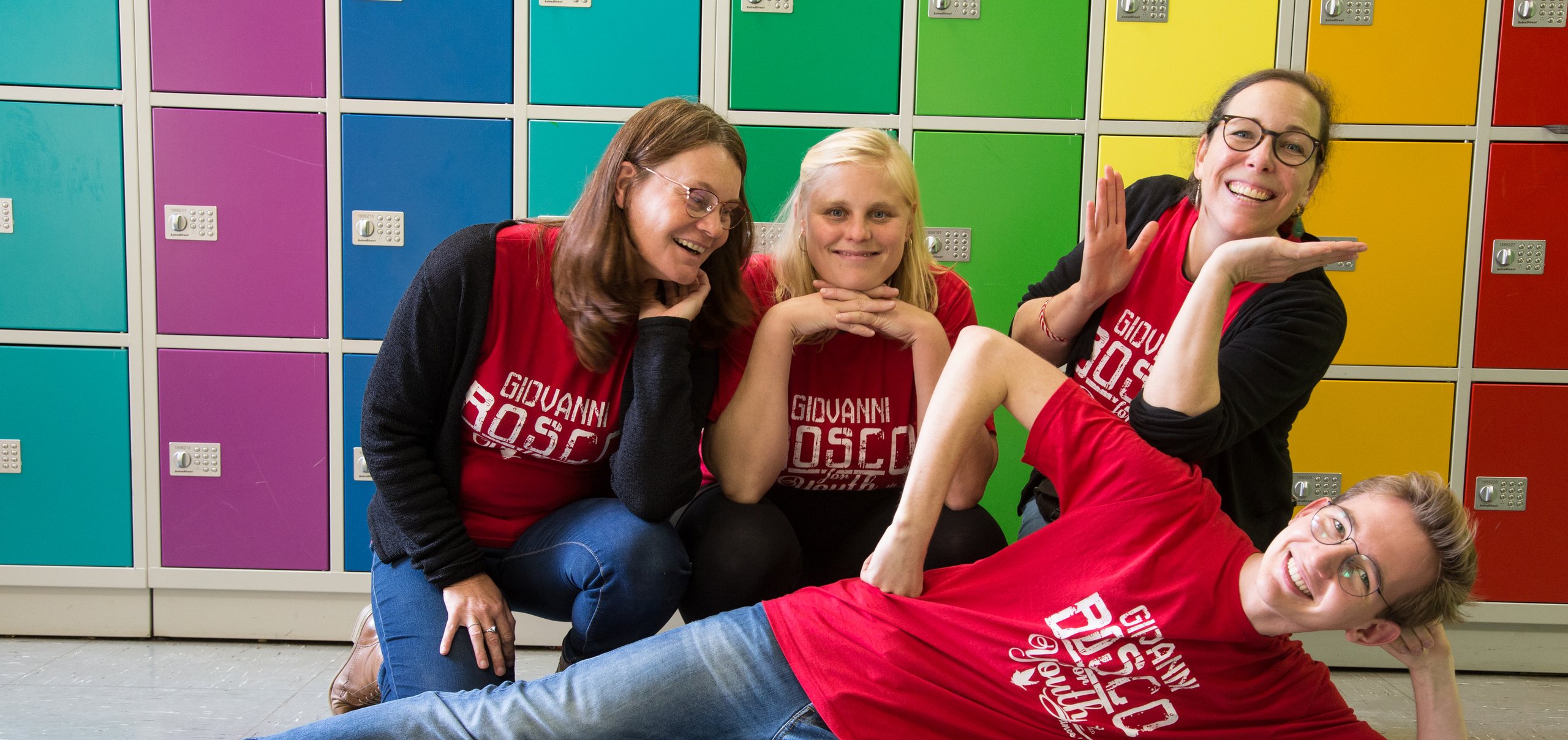 Vier lächelnde Personen, drei Frauen und ein Mann, in roten Hemden posieren spielerisch vor bunten Spinden.