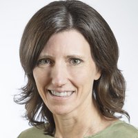 Porträt einer lächelnden Frau mit braunen Haaren und grünen Augen, die ein grünes Oberteil trägt.