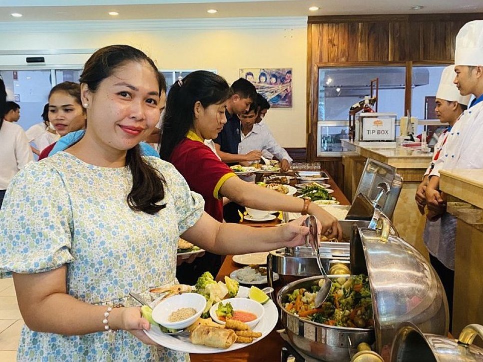 Lächelnde Frau mit vollem Teller an einem geschäftigen Buffet, mit anderen Gästen und Köchen sichtbar.