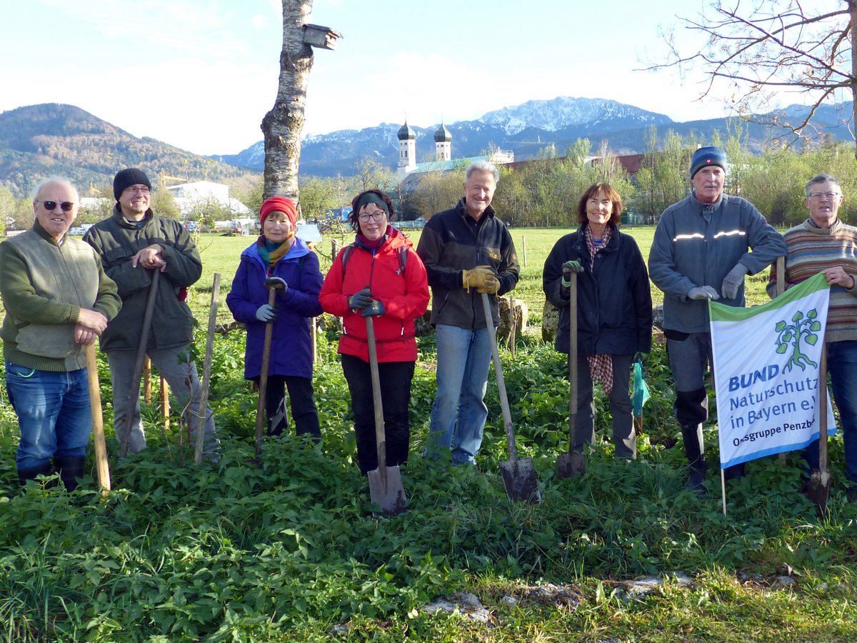 Eine Gruppe von 10 BUND Naturschutz Freiwilligen mit Spaten auf einem Feld, mit Bergen und einem Kloster im Hintergrund.