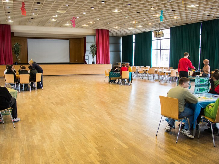 Verschiedene Gruppen von Schülern und Erwachsenen spielen Brettspiele und nehmen an Aktivitäten in einer hellen Halle teil.