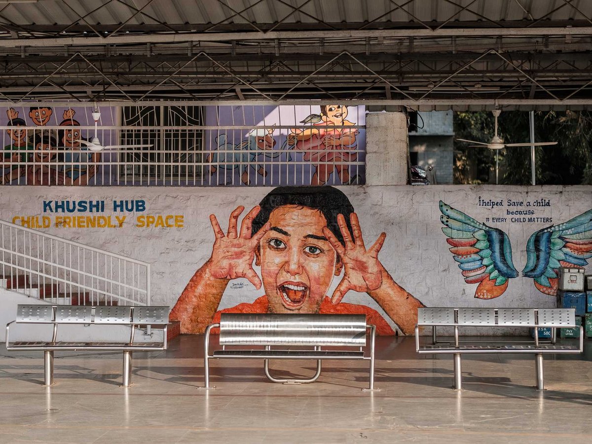 Wandgemälde eines fröhlichen Kindergesichts, bunter Flügel und des Textes „Khushi Hub Child Friendly Space“ an einer Wand mit Bänken.
