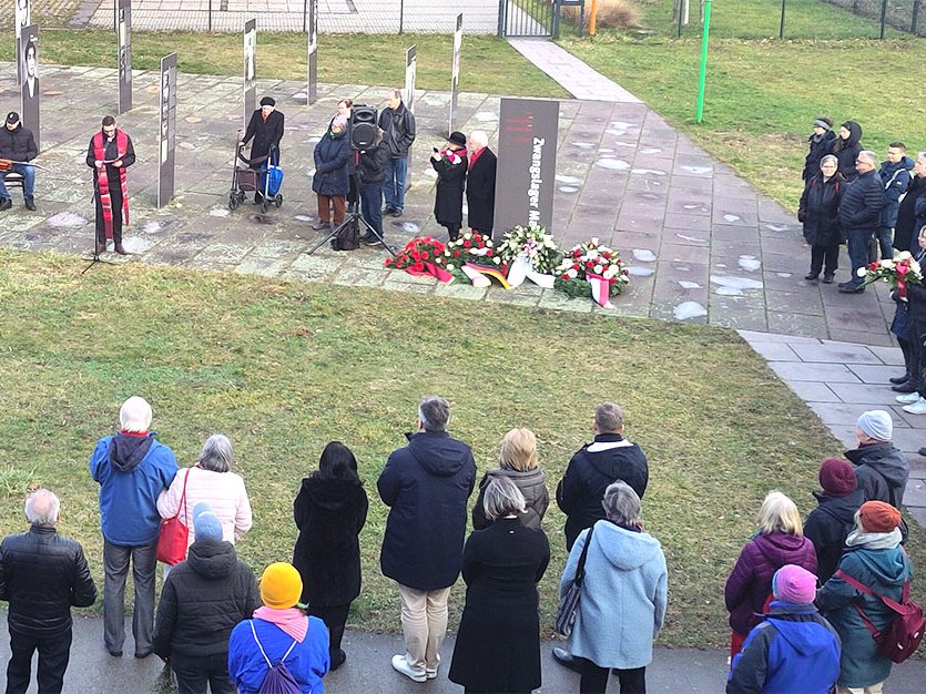 Gedenkfeier im Freien vor einem Gedenkstein mit der Aufschrift „Zwangslager“, mit Redner, Musikern, Kränzen und vielen Anwesenden.