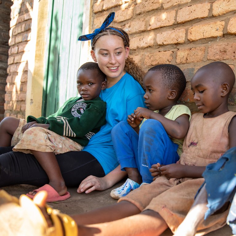 Eine lächelnde junge weiße Frau mit geflochtenen Haaren sitzt mit vier afrikanischen Kindern auf dem Boden vor einer Ziegelwand.