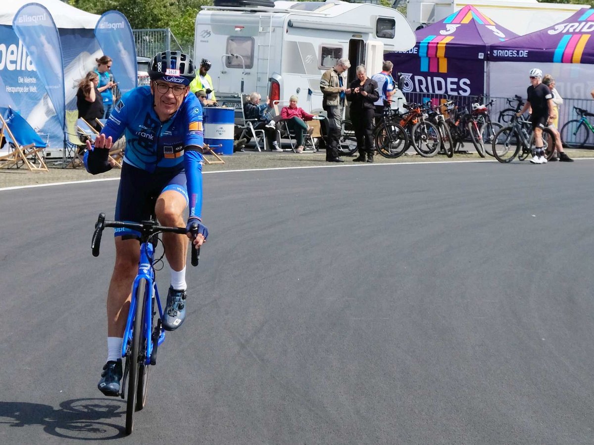 Ein lächelnder Radfahrer in blauem Trikot und Helm winkt, während er auf einem blauen Rennrad auf einer asphaltierten Strecke fährt.