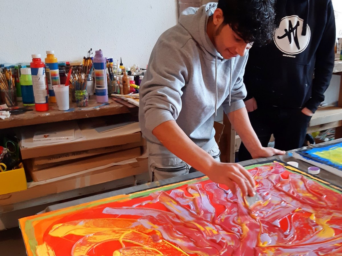 Ein junger Künstler bemalt ein abstraktes Werk mit dicken roten, gelben und lila Farben, während ein Freund im Atelier zusieht.