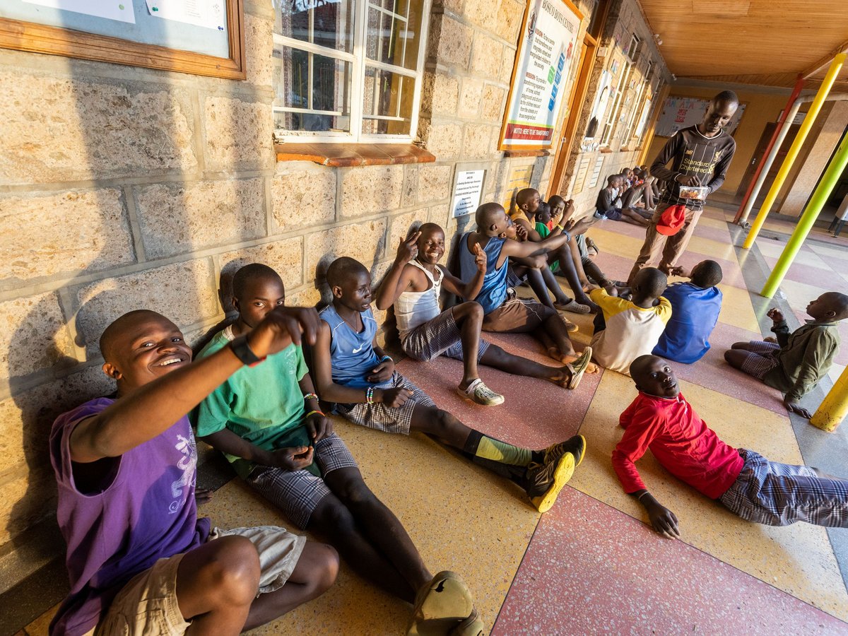 Eine Gruppe fröhlicher Jungen sitzt am Boden entlang einer Steinwand, lächelt und interagiert in einem Jugendzentrum.