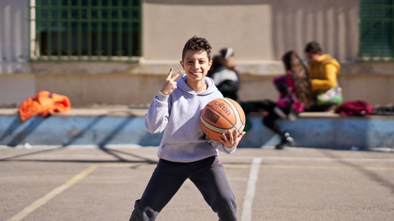 Lächelnder Junge in grauem Hoodie hält Basketball, macht Friedenszeichen, auf Außenplatz mit unscharfem Hintergrund.