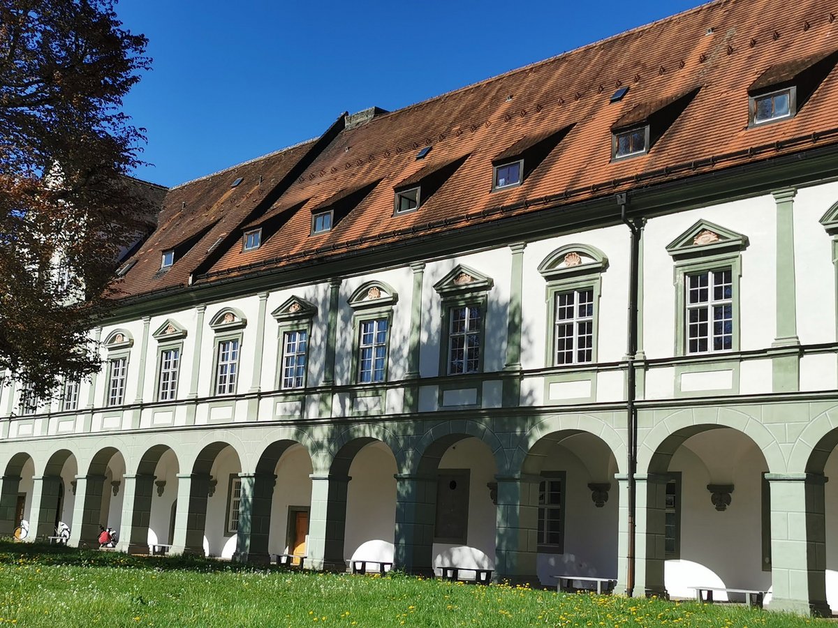 Sonnige Ansicht eines historischen Gebäudes mit langem Arkadengang, weißer Fassade, grünen Akzenten, rotem Dach, Baum und Rasen.