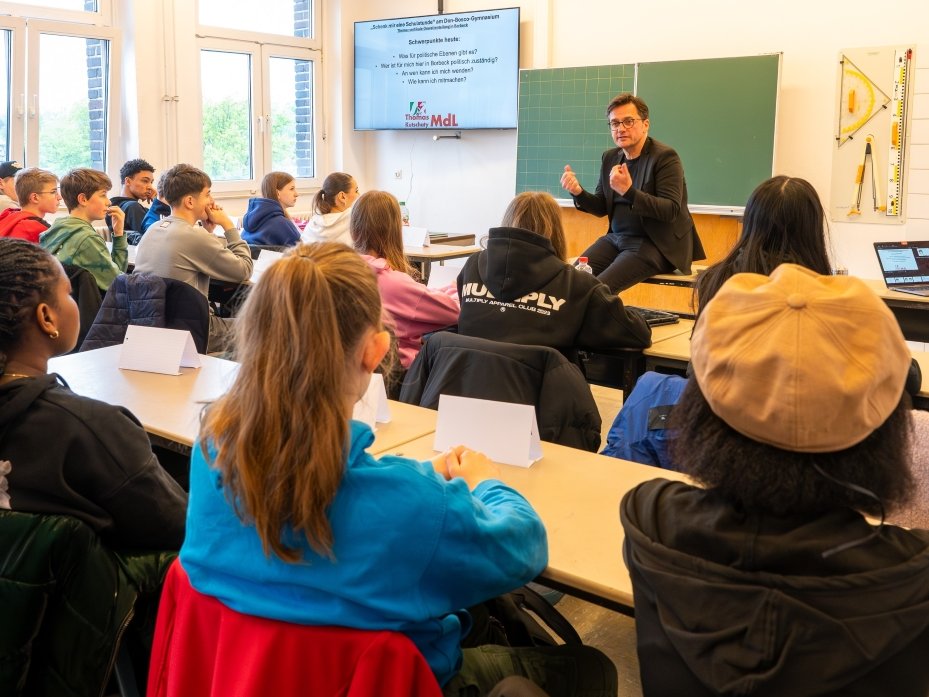 Ein Politiker spricht in einem hellen Klassenzimmer während einer Präsentation zu einer vielfältigen Gruppe von Schülern.