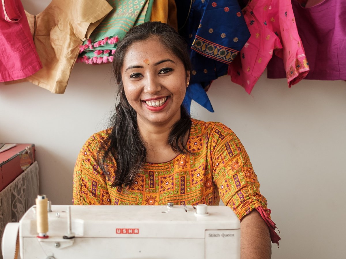 Lachende indische Frau näht an einer USHA-Maschine, umgeben von bunten, reich bestickten indischen Kleidungsstücken.