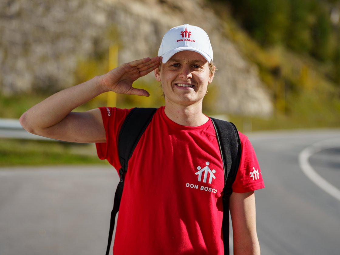 Ein lächelnder Mann in rotem Don Bosco T-Shirt und weißer Mütze salutiert, mit Rucksack und verschwommenem Straßen-/Hügelhintergrund.