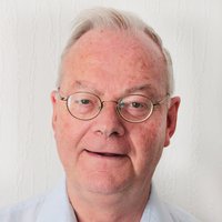 Porträtaufnahme eines älteren Mannes mit grauem Haar und Brille, der ein hellblaues Hemd trägt und leicht lächelt.