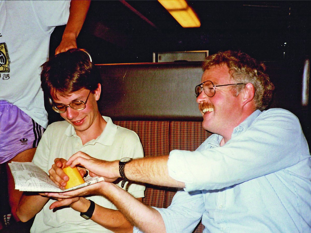 Ein lächelnder Mann mit Schnurrbart bietet einem jüngeren Mann Käse an, beide tragen Brillen, in einem Zug oder Bus.