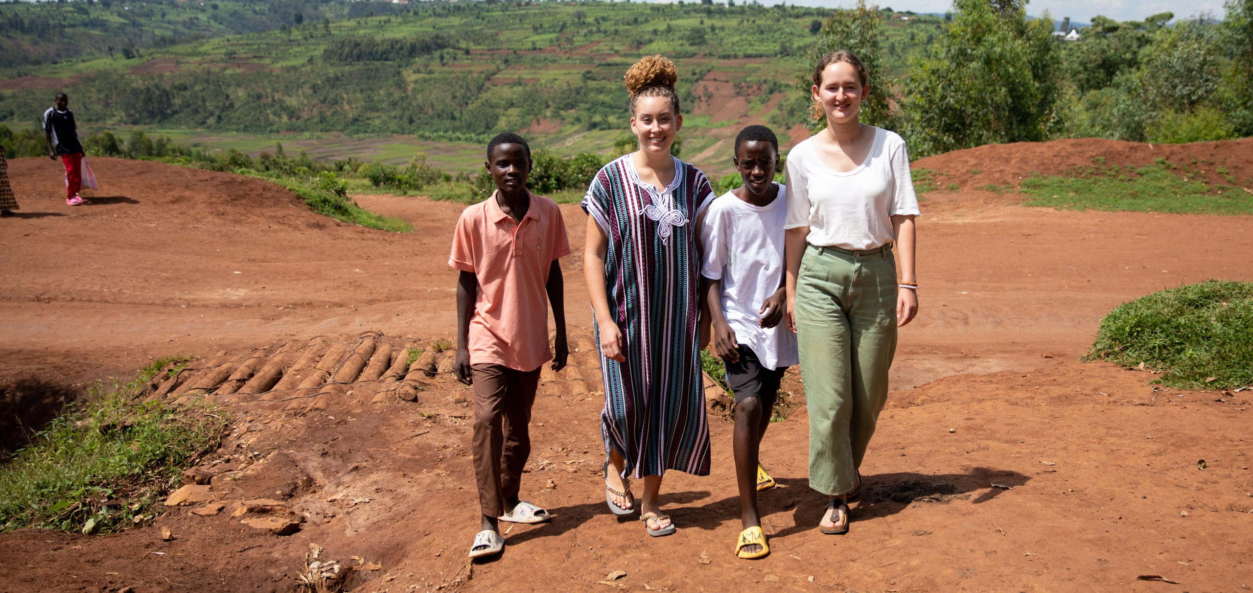 Zwei Jungen und zwei Frauen gehen auf einem unbefestigten roten Weg in einer hügeligen, grünen Landschaft unter bewölktem Himmel.