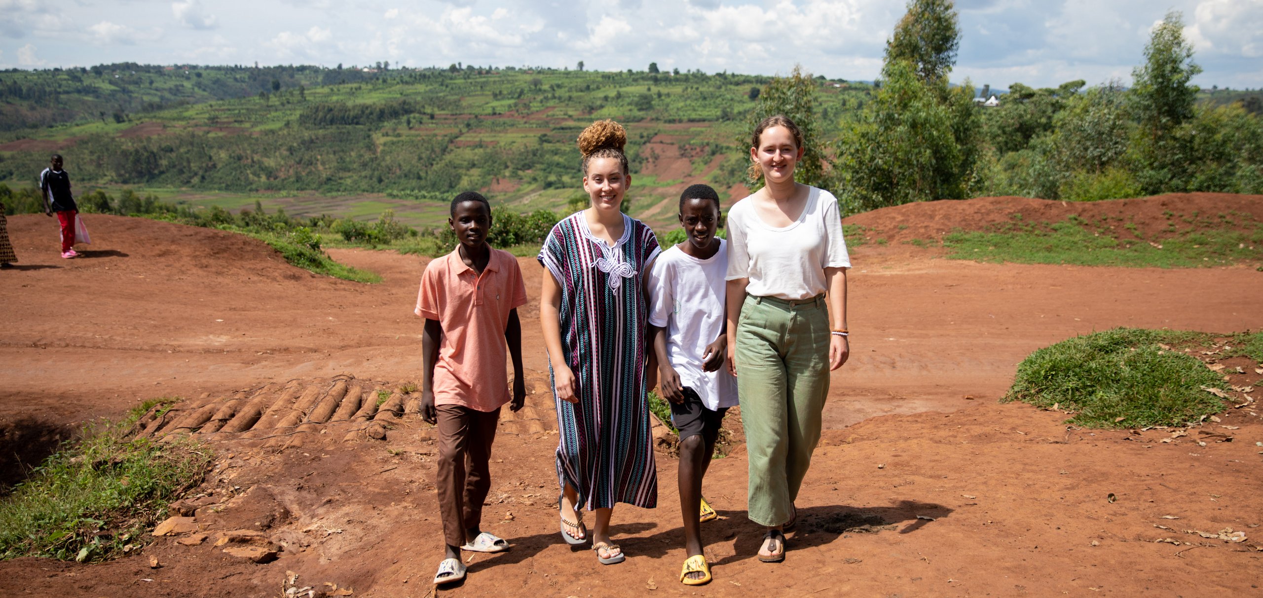 Zwei Jungen und zwei Frauen gehen auf einem unbefestigten roten Weg in einer hügeligen, grünen Landschaft unter bewölktem Himmel.
