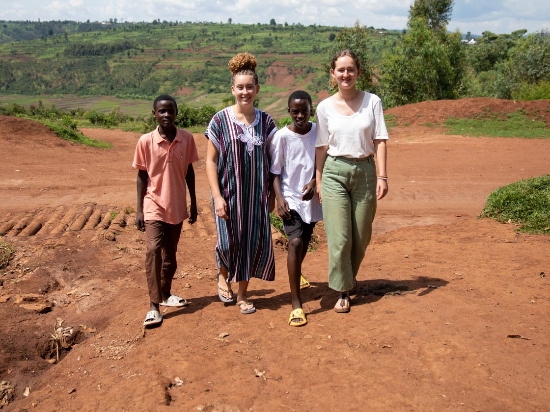 Zwei Jungen und zwei Frauen gehen auf einem unbefestigten roten Weg in einer hügeligen, grünen Landschaft unter bewölktem Himmel.