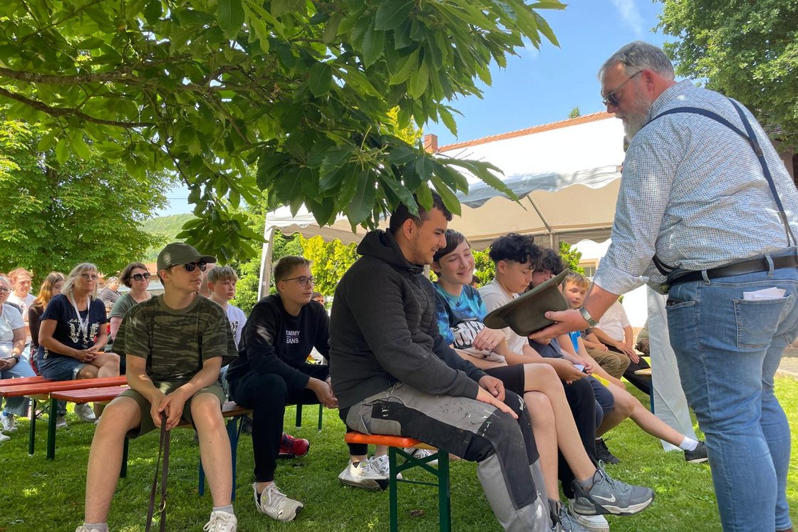 Ein bärtiger Mann mit Hosenträgern überreicht sitzenden Jugendlichen bei einer Veranstaltung im Freien unter einem Baum einen Hut.