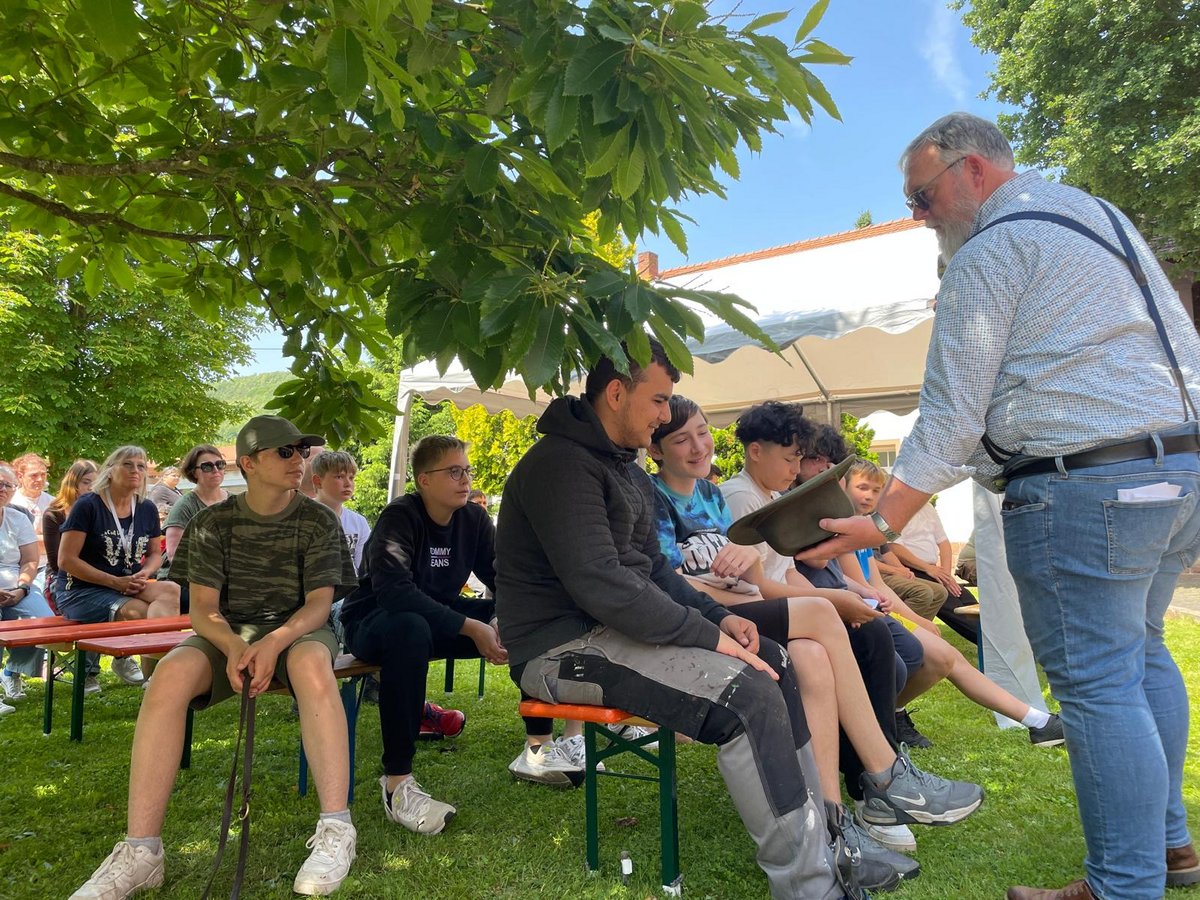 Ein bärtiger Mann mit Hosenträgern überreicht sitzenden Jugendlichen bei einer Veranstaltung im Freien unter einem Baum einen Hut.
