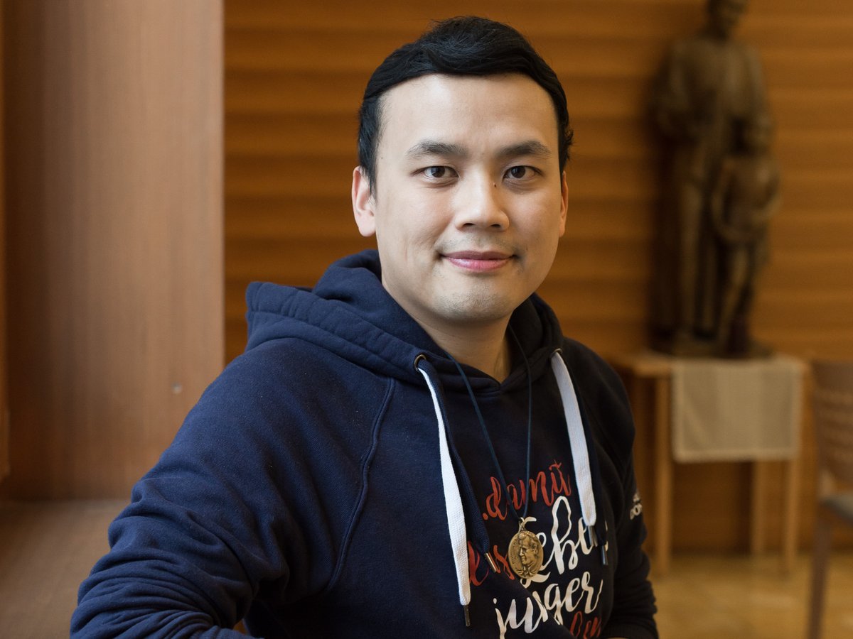 Ein Mann ostasiatischer Herkunft in dunkelblauem Kapuzenpullover und goldenem Medaillon, sitzend und lächelnd.