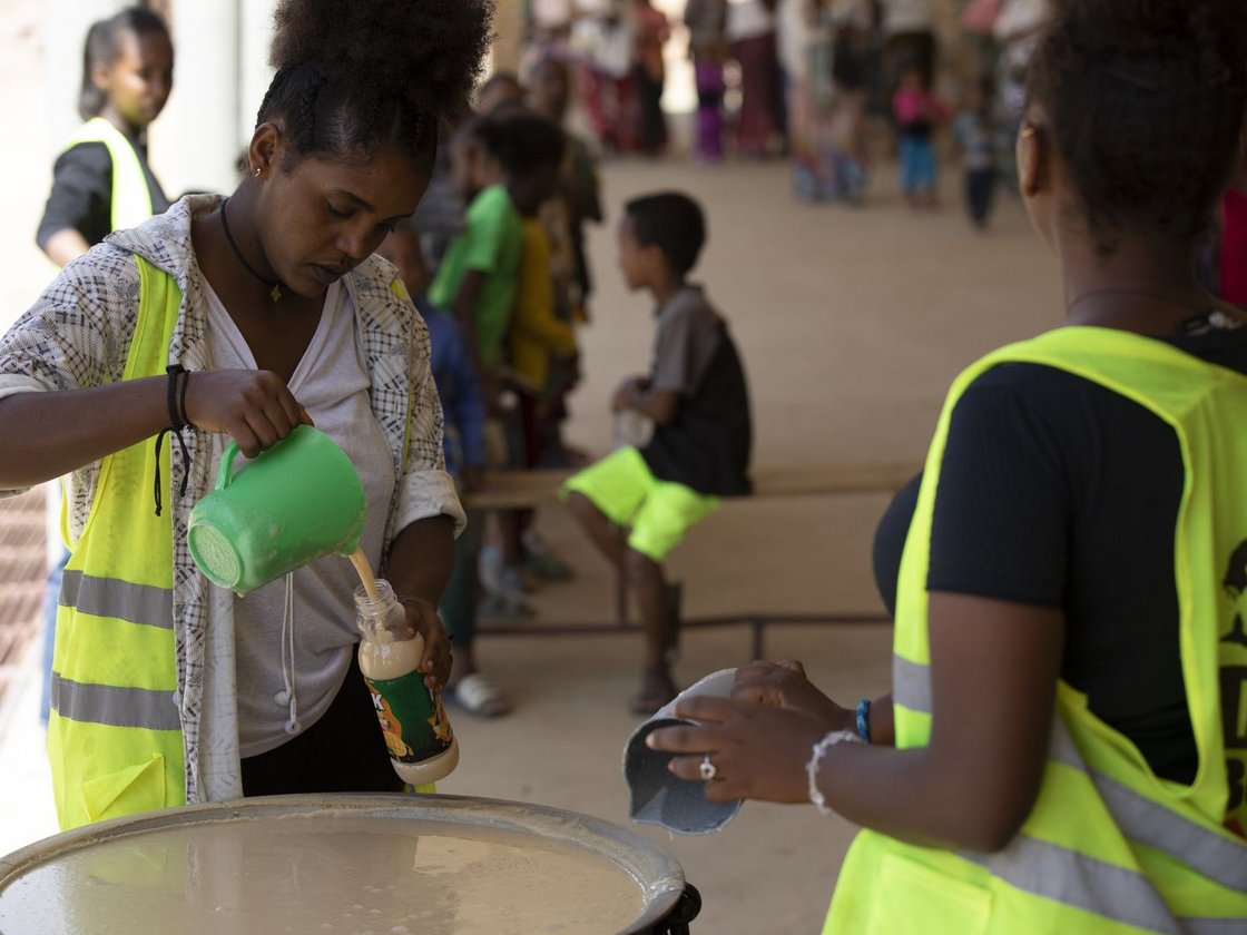 Zwei Frauen in Warnwesten verteilen Getränke aus großem Behälter, füllen Flaschen für Menschen im Hintergrund.