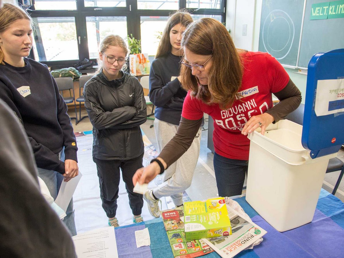 Eine Frau zeigt Schülern, wie man Papier in einem Behälter entsorgt, im Rahmen einer 'Welt Klima Challenge' Präsentation.