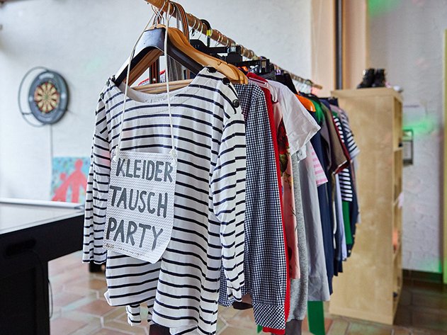 Ein Kleiderständer mit verschiedenen Kleidungsstücken und einem Schild 'KLEIDER TAUSCH PARTY' an einem Ringelshirt. Im Hintergrund ist eine Dartscheibe.