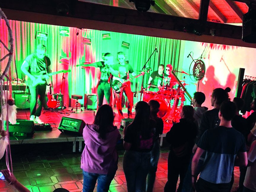 Eine Live-Band spielt auf einer Bühne unter leuchtenden roten und blauen Lichtern, während ein Publikum zuschaut.