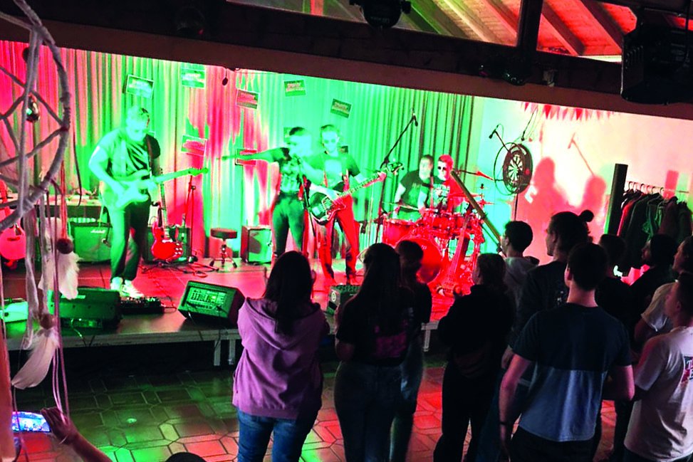 Eine Live-Band spielt auf einer Bühne unter leuchtenden roten und blauen Lichtern, während ein Publikum zuschaut.