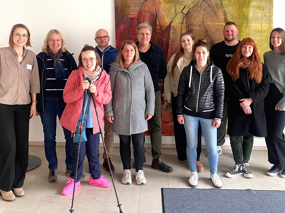 Eine Gruppe von 11 lächelnden Personen, Männer und Frauen, stehen drinnen vor einem abstrakten Gemälde. Eine Frau hält Trekkingstöcke.