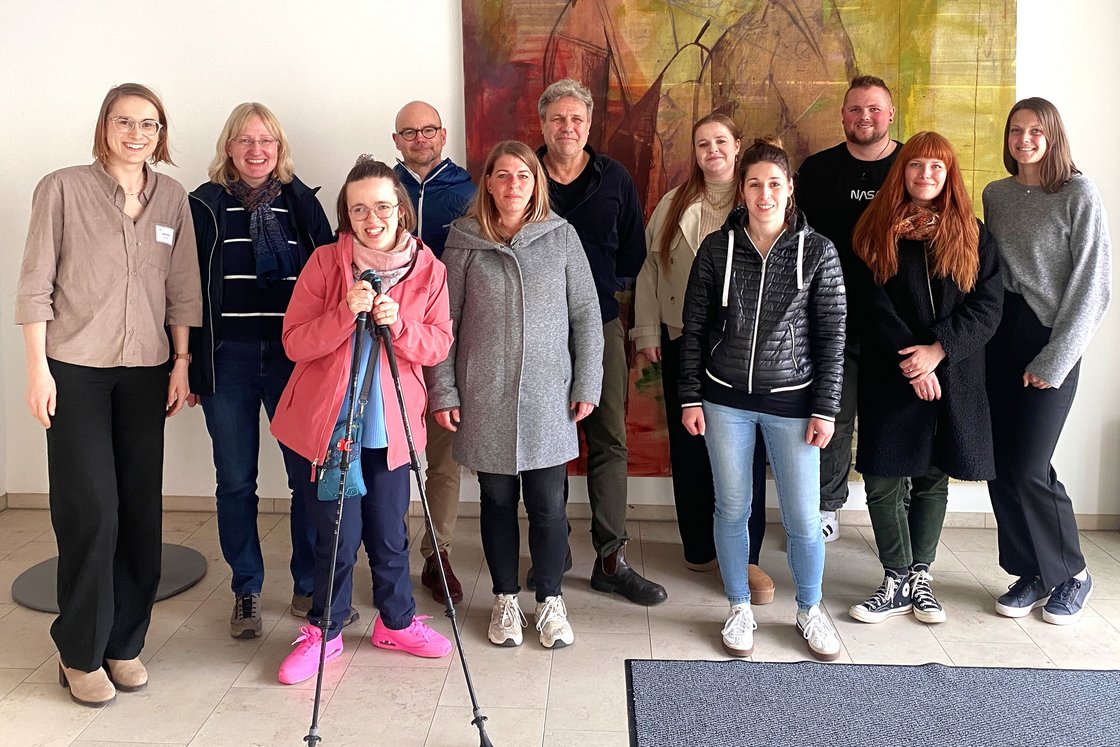 Eine Gruppe von 11 lächelnden Personen, Männer und Frauen, stehen drinnen vor einem abstrakten Gemälde. Eine Frau hält Trekkingstöcke.