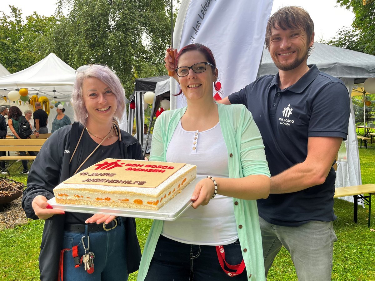 Drei lachende Personen halten eine Torte zum 10-jährigen Jubiläum von Don Bosco Sachsen Jugendhilfe bei einem Outdoor-Event.