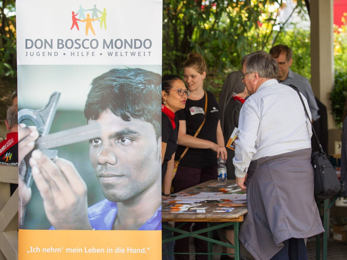 Ein Infostand von Don Bosco Mondo mit einem Banner, das einen jungen Mann bei der Arbeit zeigt. Menschen betrachten Broschüren.