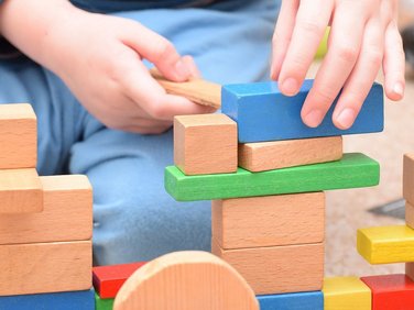 Kinderhände bauen mit bunten Holzklötzen. Blaue, grüne, naturfarbene, rote und gelbe Klötze sind gestapelt.
