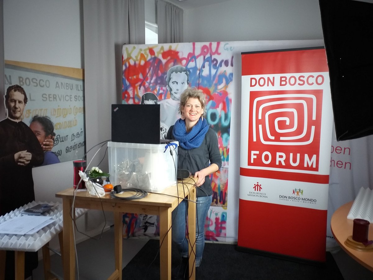 Lächelnde Frau mit blauem Schal hinter Tisch bei Don Bosco Forum-Veranstaltung mit Bannern und Graffiti-Kunst.