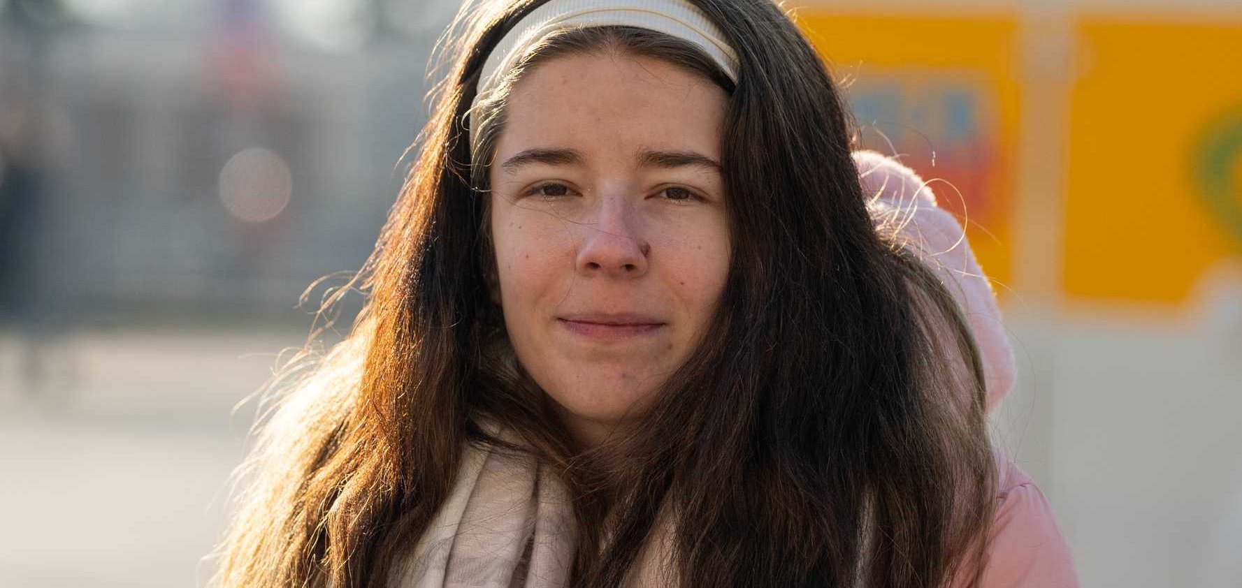 Junge Frau mit langen dunklen Haaren, weißem Stirnband, rosa Jacke und Schal blickt nach vorne im Freien.