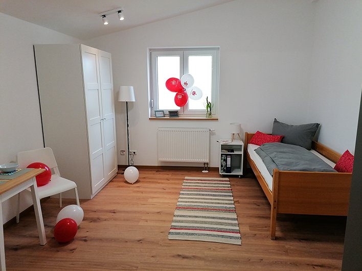 Helles, modernes Zimmer mit Holzfußboden, Einzelbett, weißem Kleiderschrank, Tisch und rot-weißen Ballons am Fenster.