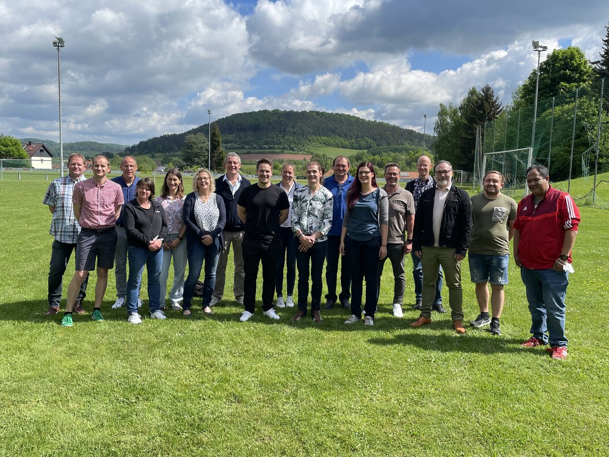 Eine Gruppe von 19 lächelnden Menschen steht auf einem Sportplatz vor einer bewaldeten Hügellandschaft unter einem Himmel mit weißen Wolken.